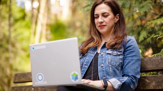 Ashely Rajagopal坐在外面的长凳上,身后是生机勃勃的绿色植物。她在笔记本电脑上工作时很专注。