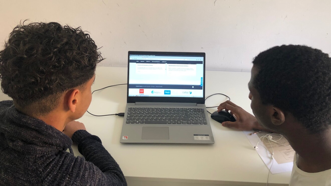 La donación de Amazon a la Fundación Diagrama ha sido de 39 ordenadores portátiles y 72 tabletas. En la foto aparecen dos chicos negros en primer plano con el peli también negro. Unos lleva una camisa negra y el otro una camiseta blanca. Entre ellos hay un ordenador portátil encendido y el chico de la derecha sujeta el ratón con la mano. La mesa es de color blanco y la pared también.
