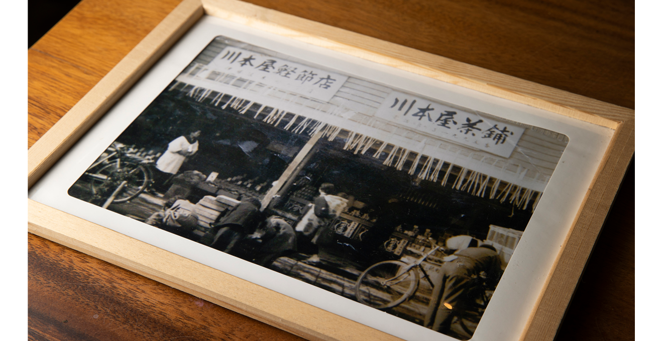 古い白黒写真。川本屋鰹節店と川本屋茶舖の二つの看板を掲げた店舗に割烹着を着た女性や子どもをおんぶした女性、自転車で来た人たちが映っている。