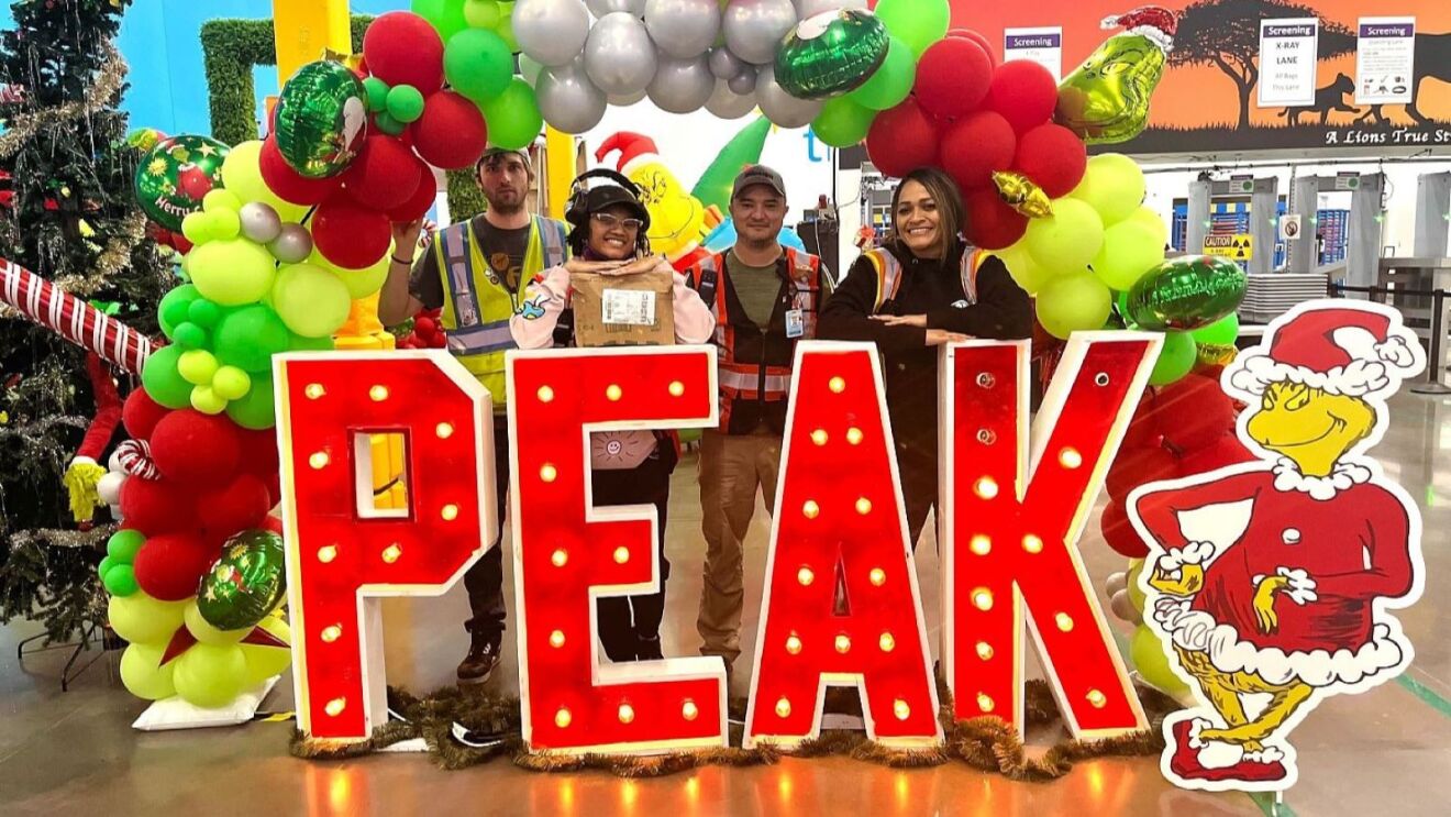 Four workers pose behind large 'PEAK' sign, surrounded by festive decorations and Grinch character