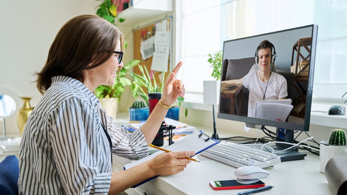 A woman wearing glasses sits at a desk and video conferences with a man wearing headphones and holding up a notebook.