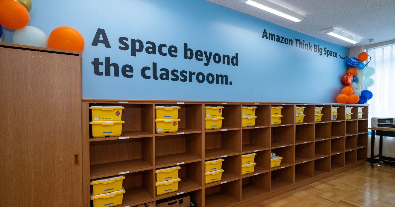教室後方の壁と棚。壁は水色に塗られ、「A space beyond the classroom」と「Amazon Think Big Space」という黒い文字。壁の下側の茶色い木目調の棚の最上段と三段目、下段一部に黄色い収納ボックスが重ねて収納されている。壁の両側はオレンジ、白、青の風船で飾られている。