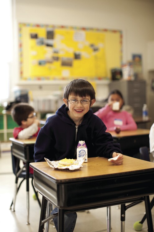 A boy sits at a school desk, smiling at the camera. On his desk are a carton of milk and a breakfast sandwich. Behind him, other students also eat and drink.