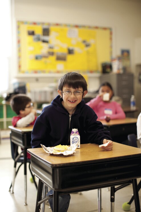 A boy sits at a school desk, smiling at the camera. On his desk are a carton of milk and a breakfast sandwich. Behind him, other students also eat and drink. 