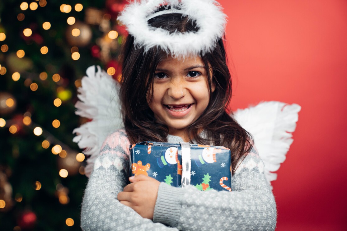 Ein dunkelhaariges Mädchen mit weißen Engelsflügeln hält ein blaues Geschenk in den Armen und lächelt in die Kamera. Im Hintergrund sieht man einen geschmückten Weihnachtsbaum.