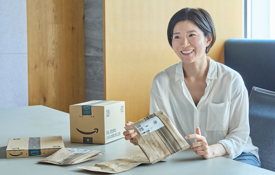 オフィスの室内で広いデスクに向かって座る女性。手に梱包資材を持っている。デスクの上にも複数の箱や袋。
