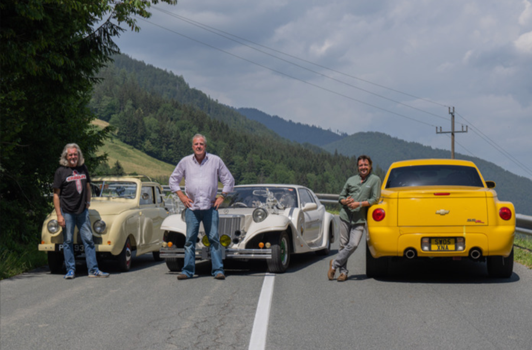 Three men (Jeremy Clarkson, Richard Hammond, and James May) pose in the middle of the road in central Europe behind three cars