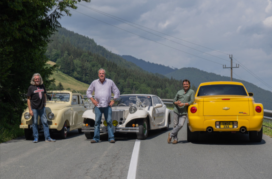 Three men (Jeremy Clarkson, Richard Hammond, and James May) pose in the middle of the road in central Europe behind three cars 