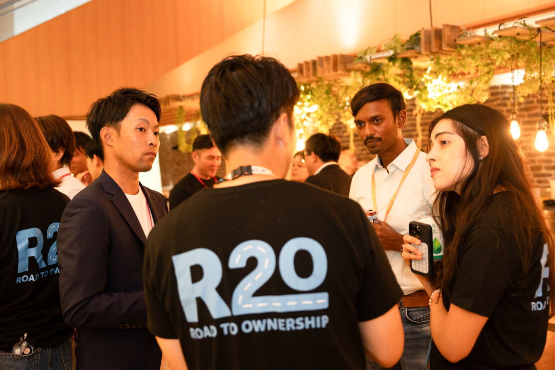 後ろにR2Oという青文字で印刷されているTシャツを着ている男性が同じTシャツを着ている女性、白いシャツを着ている男性とジャケットを着ている男性と話しているようだ。みんなが彼に注目されている。