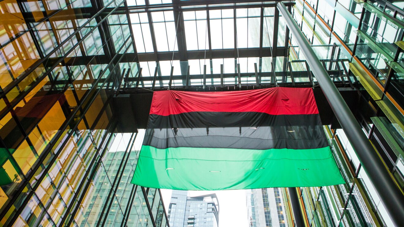 A flag with red, black, and green horizontal stripes flies from a skybridge between two office buildings.