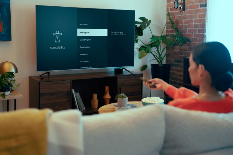 An image of a woman watching television on her couch. She is wearing a hearing aid in one ear and selecting an accessibiliyt option on her Fire TV device.