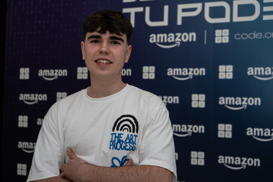 Con un fondo de photocall con el logo de Amazon y de Code. org, en blanco sobre un fondo azul aparece unos de los finalistas, Hugo Centeno Sanz. Está con los brazos cruzados. Lleva una camiseta de manga corta con el texto The art process.