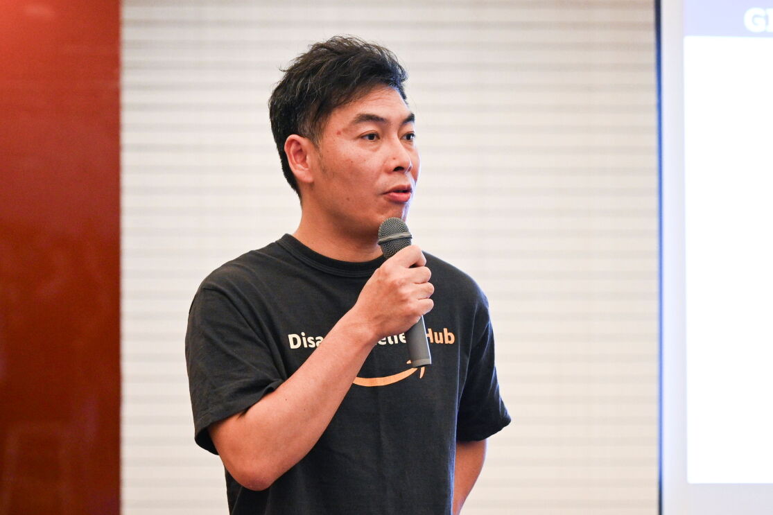 「Disaster Relief Hub」のロゴが入った黒いTシャツを着ている男性（瀧川匠さん）がマイクで話している。