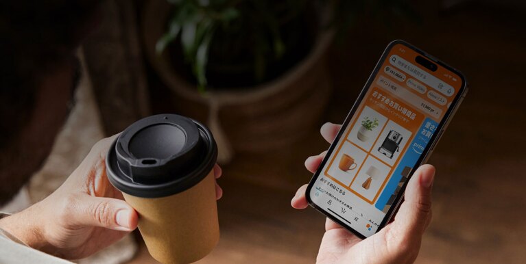 左手に紙製のテイクアウトカップ、右手にスマートホンを持つ人物の手元のショット。スマートホンの画面にはオレンジ色を基調としたAmazonアプリの「おすすめお買い得商品」が表示されている。