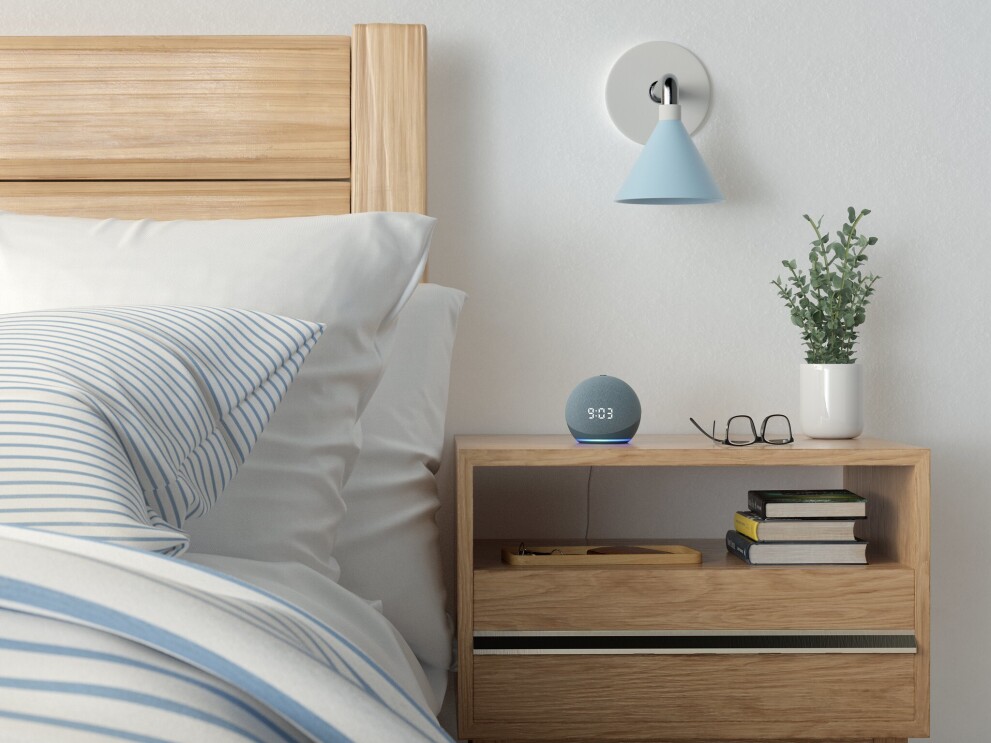 A shot of the new Echo Dot on a bedside table, there is also a plant and a pair of glasses on the table. To the left is a bed.