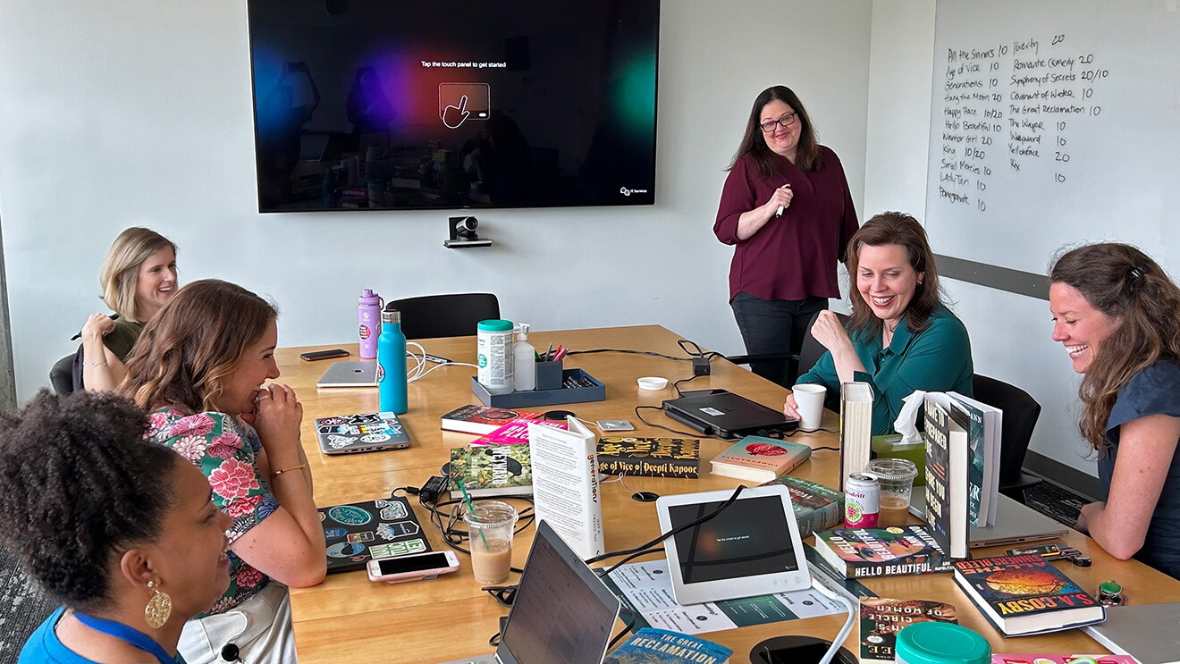 A photo of Amazon editors in a meeting room, sitting around a table that has multiple books and laptops on it, viewing a spreadsheet of notes on a screen.
