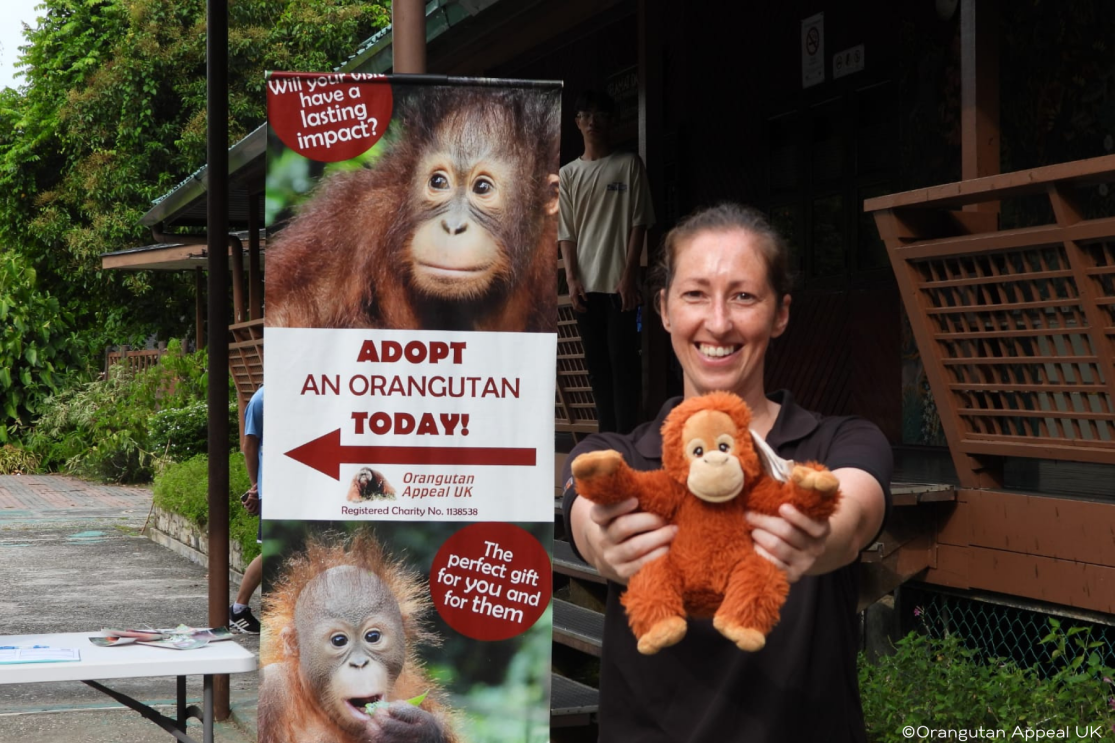 Ann Towse with a toy orangutan in front of a sign urging people to adopt an orangutan today