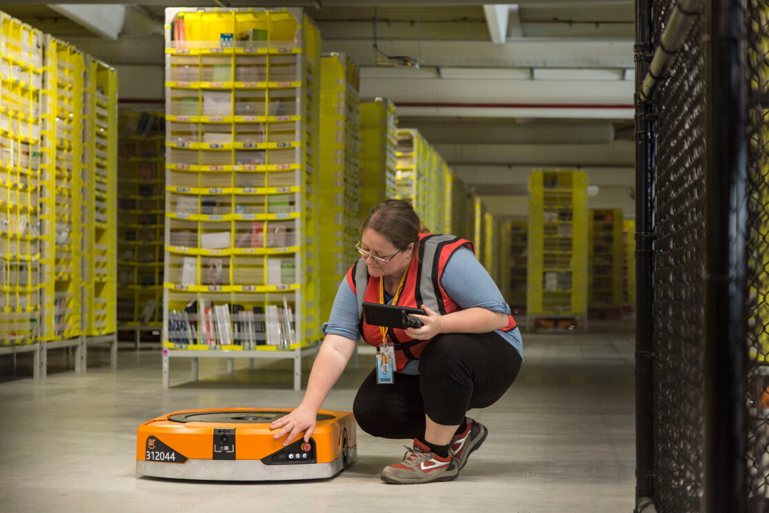 Foto di Maria Stella Carnà che regge un tablet di controllo e si china su uno dei robot di cui è responsabile all'interno del Centro di Distribuzione Amazon di Passo Corese