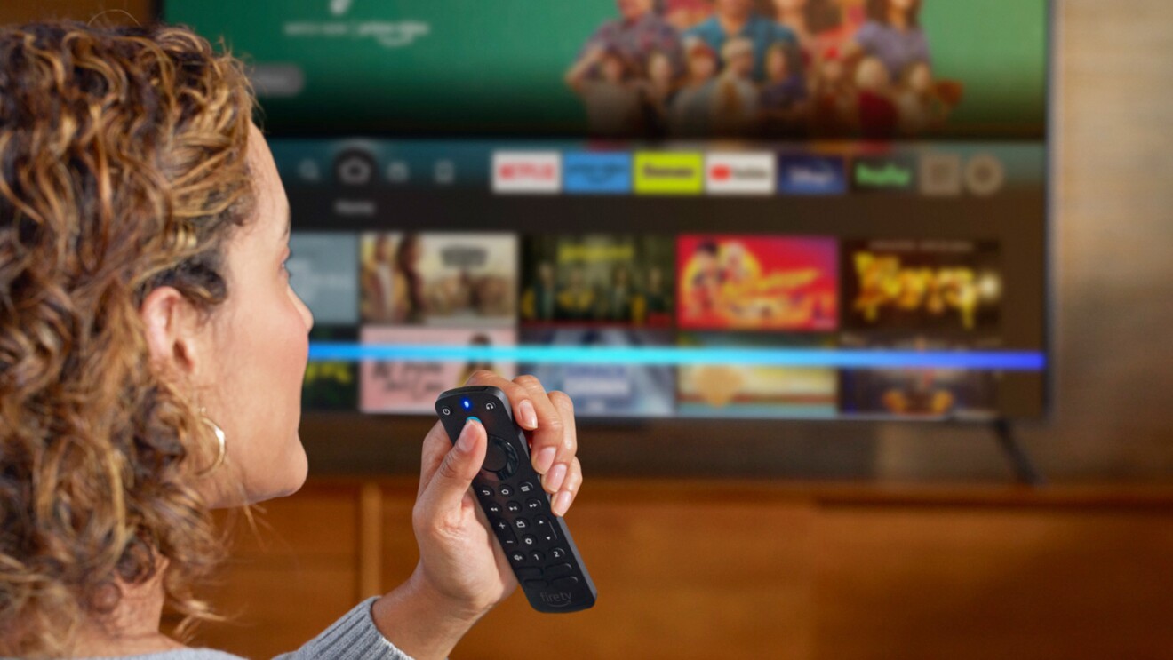 An image of a woman pressing a button on a TV remote speaking with Alexa. There is a TV blurred in the background.