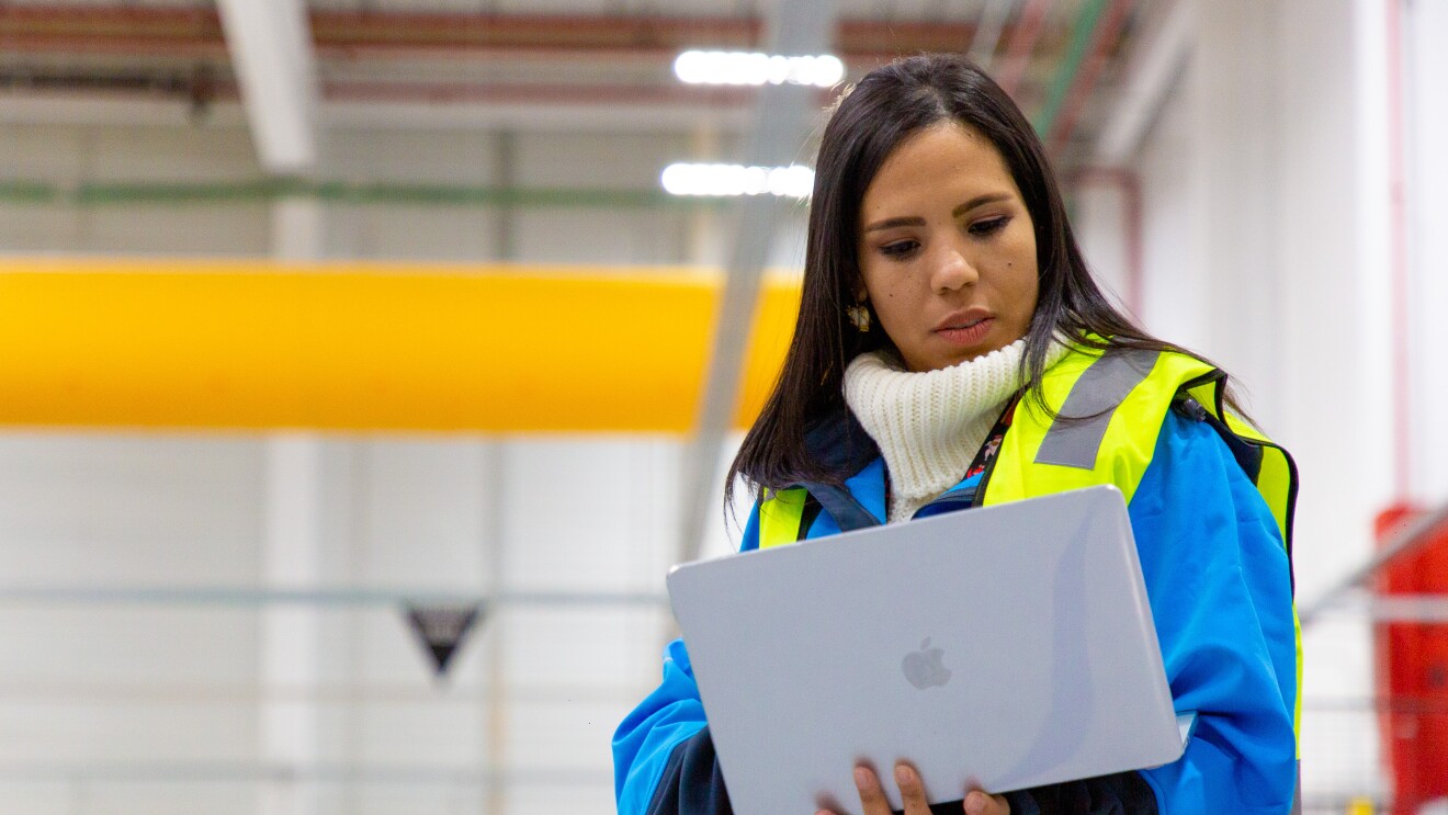 Ivette Duran, CEO de Cainaima Logistics, de pie con un ordenador entre las manos. De fondo está el centro logístico de color blanco y amarillo. Ella es morena y tiene el pelo largo y negro. Lleva un jersey de color blanco y de cuello alto, una chaqueta azul y un chaleco fluorescente. 