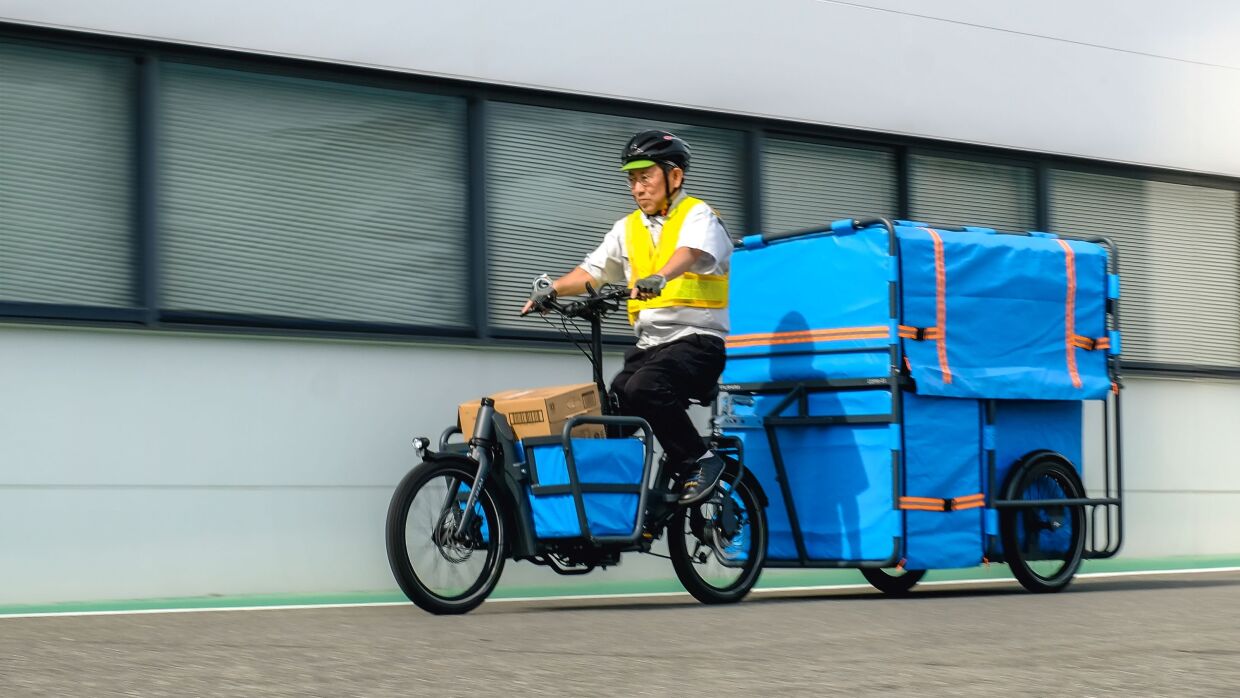 安全ベストを着ている男性が自転車に乗っている。その自転車の前に付けたカゴにダンボールが入っている。その自転車の後ろには大きな青いリヤカーが付けられている。
