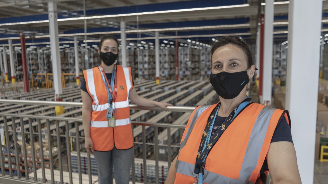 Marta Torrejón Serrano y María Jesús Serrano. Madre e hija posan en un balcón del centro logístico de Illescas que se ve de fondo. Están apoyadas a la banadilla manteniendo la distancia de seguridad. Las dos van con manga corta, mascarilla corporativa y chaleco naranja. Llevan el pelo recogido con una coleta.