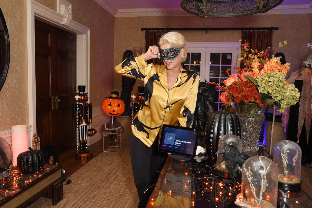 Image of a woman standing next to a table in her hallway with Halloween decorations and an Echo Show device. She is wearing a masquerade mask while smiling for the photo.