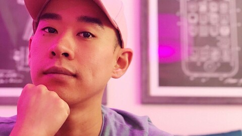 一名男子在粉色灯光下微笑拍照的大头照。他戴着一顶白色的棒球帽,下巴托在拳头上。
