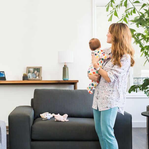 A mom holds her baby as she looks across the living room at a Echo Show device.