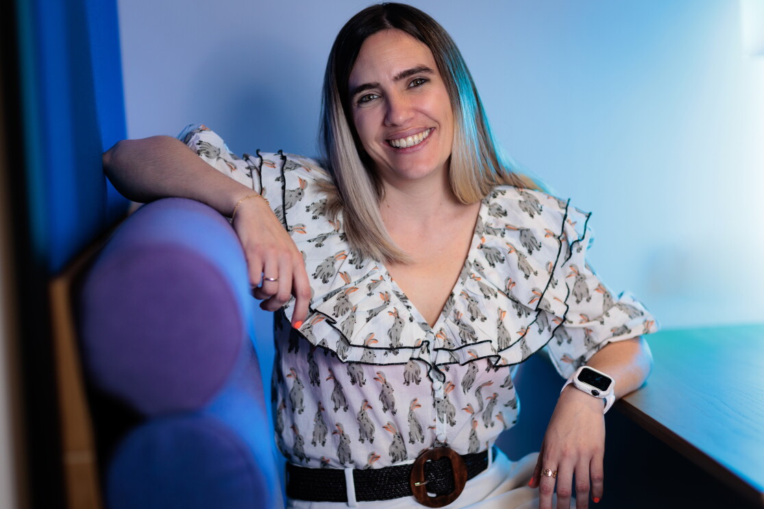 Lara Fajardo va con una camisa blanca estampada, con el pelo hasta el hombro con mechas negras y decoloradas hasta el rubio en las puntas. Con pantalones blancos y un cinturín de color negro. Con uñas rosas y un anilo en cada mano. En la mano derecha un reloj. Esta mano está apoyada en la mesa y la otra en un sofá de color azul y morado.