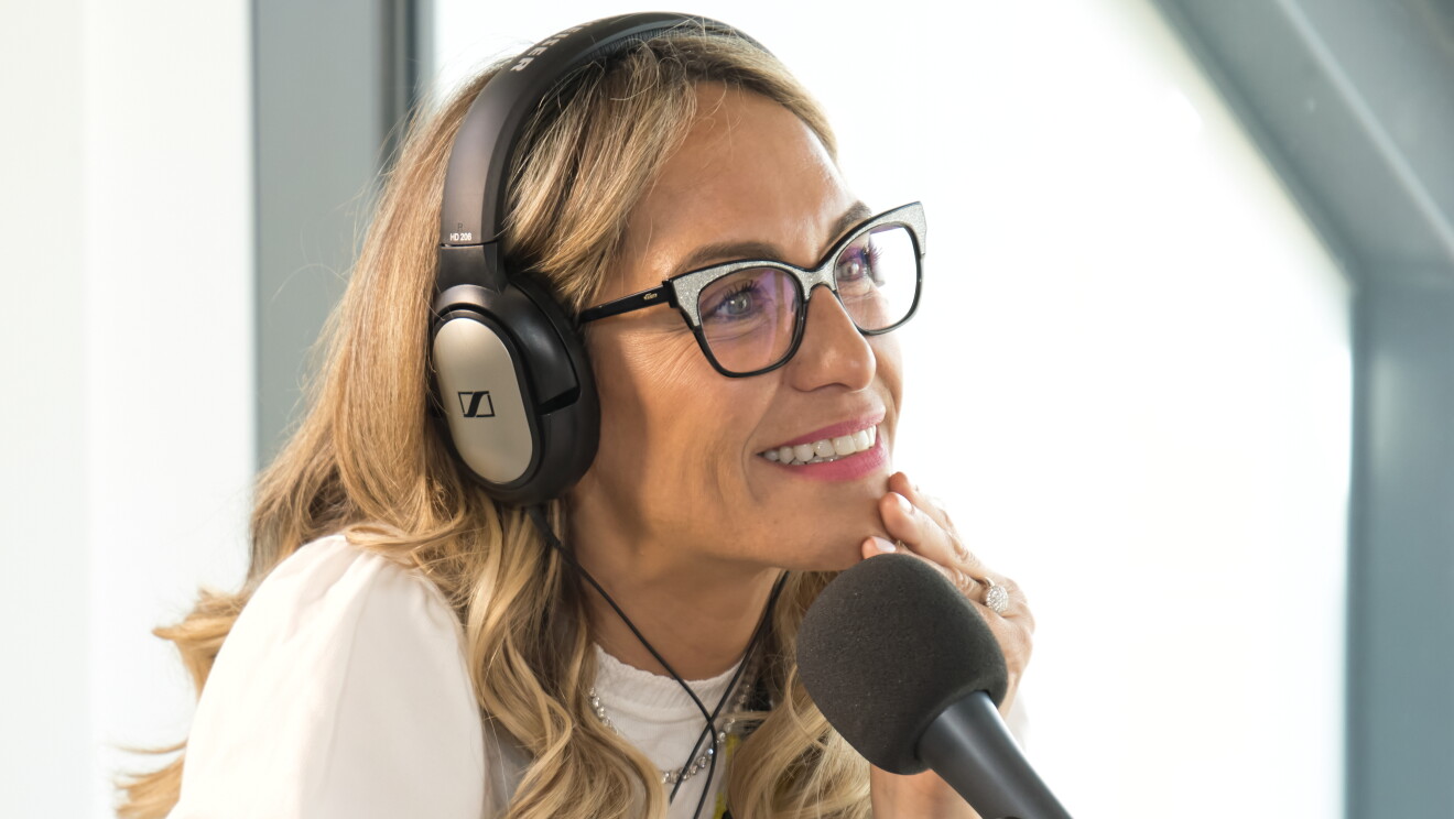 Luján Argüelles, presentadora del Podcast Despega con Amazon. Ella está mirando a un interlocutor que no se ve. Lleva el pelo suelto por el hombro  y color rubio. Una jresey color blanco con una flor de color amarillo, un collar negro, los casco puestos y una gafas de montura de pasta negras y plateadas. 