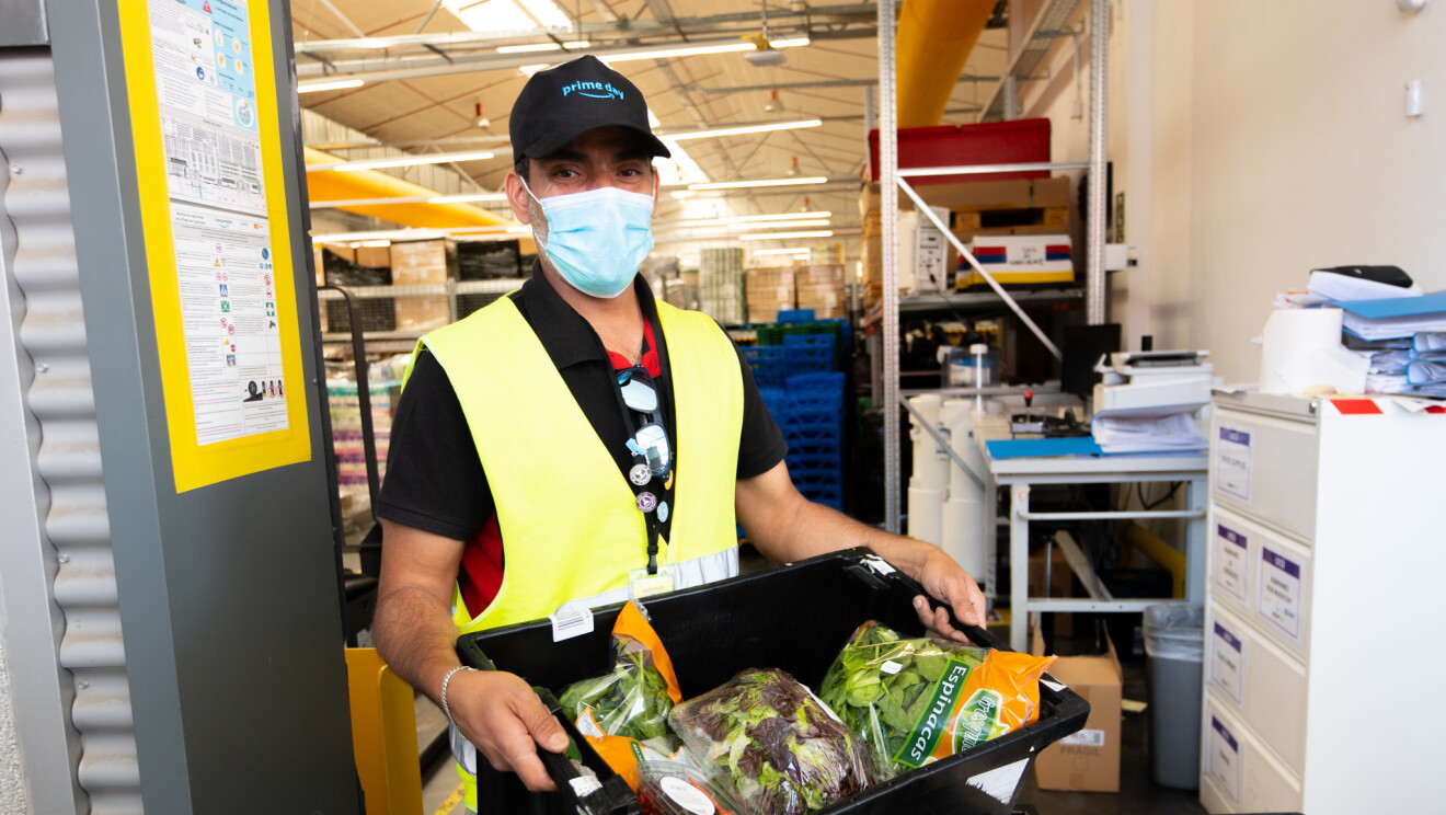 En un centro logístico de Amazon hay un trabajador con pantalones deportivos de color gris, camiseta negra de manga corta y chaleco de color amarillo. Lleva mascarilla quirúrgica y una gorra negra con el lema de Prime Day. Está agarrando una caja negra de comida, ensaladas en bolsa. De fondo el interior de un centro logísticco con estanterías llenas de productos . 