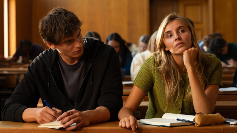 Man dressed in black looks at the girl sitting next to him in a lecture theatre as she looks forwards