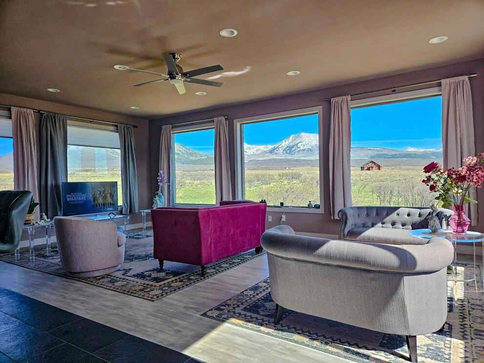 An image of a living room with mauve and pink furnishings. There are floor-to-ceiling windows surrounding the room showing views of green fields and the snow-capped Rocky Mountains.