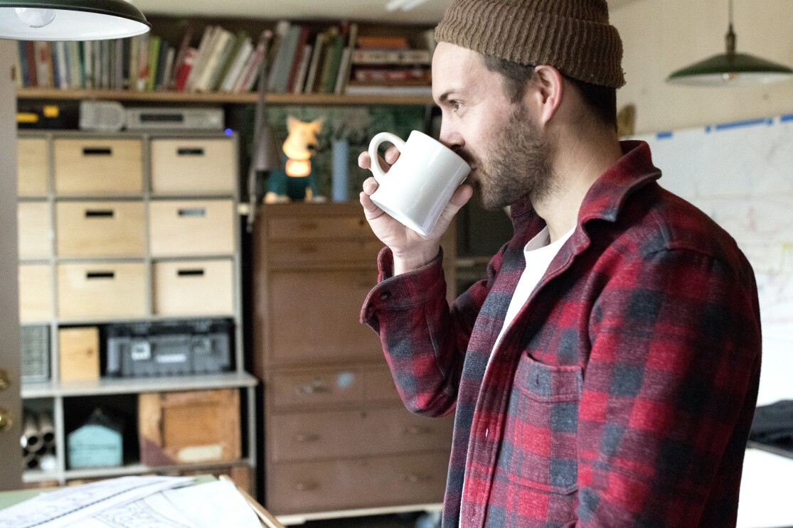 Kyler Martz in his Seattle studio, drinking a cup of coffee. He is wearing a brown hat and a red and black checked flannel shirt.