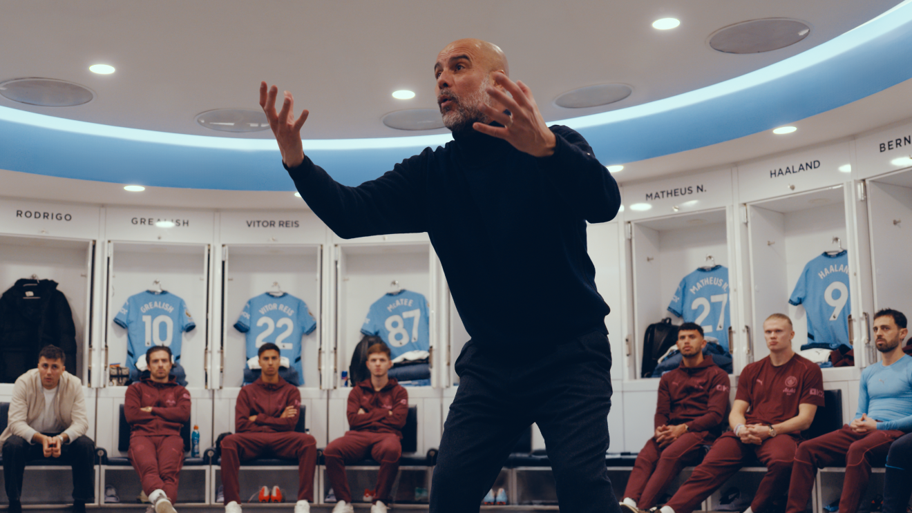 Pep Guardiola addresses his players in the middle of dressing room, arms in the air