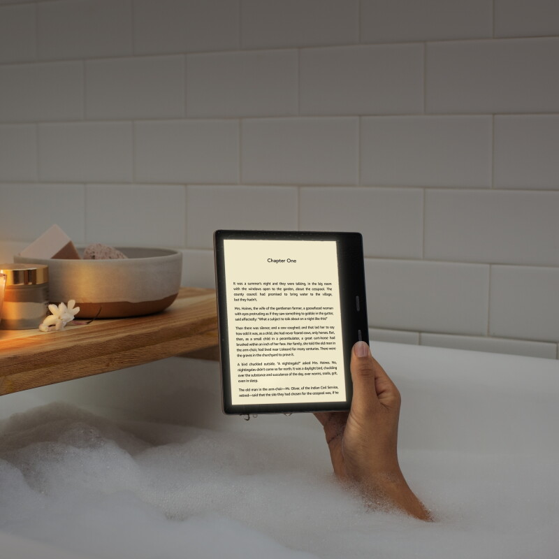 A woman holds a Kindle Oasis device with the page of a book on the display. Her arm appears from bubbles in a tub, with a wooden tub caddy behind the device.