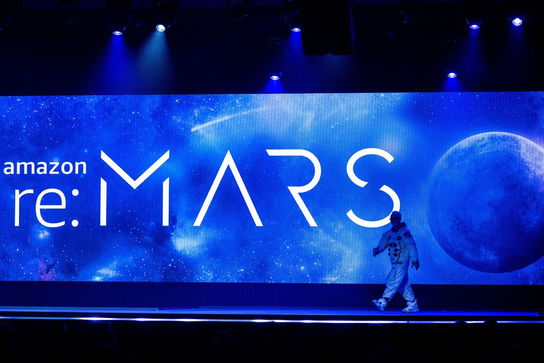 An image of the stage at re:MARS 2022. A man is walking onto the stage in an astronaut suit. In the background there is a large screen that says "Amazon re:MARS" with an image of a moon and space.