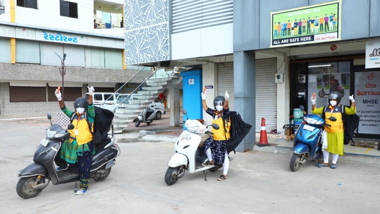 三名骑着轻便摩托车的妇女穿着黄色的亚马逊背心,背上背着快递袋,都竖起了两个大拇指。