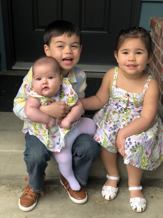 An image of two children smiling for a photo on a doorstep. One child is holding a baby and they appear to be siblings dressed in color-coordinated outfits.