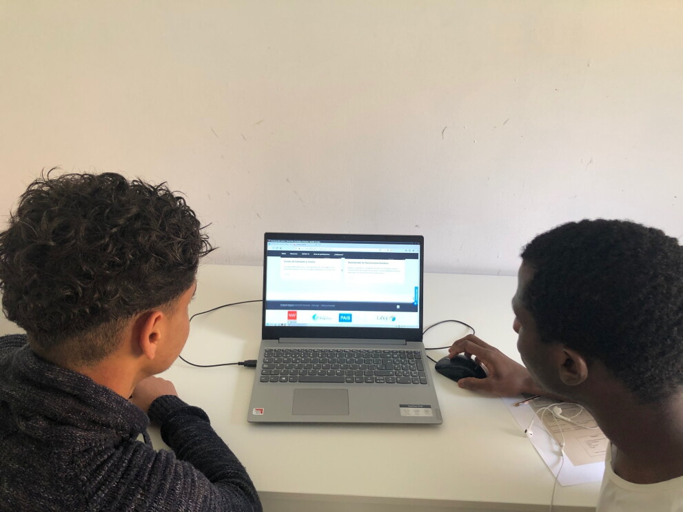 La donación de Amazon a la Fundación Diagrama ha sido de 39 ordenadores portátiles y 72 tabletas. En la foto aparecen dos chicos negros en primer plano con el peli también negro. Unos lleva una camisa negra y el otro una camiseta blanca. Entre ellos hay un ordenador portátil encendido y el chico de la derecha sujeta el ratón con la mano. La mesa es de color blanco y la pared también. 