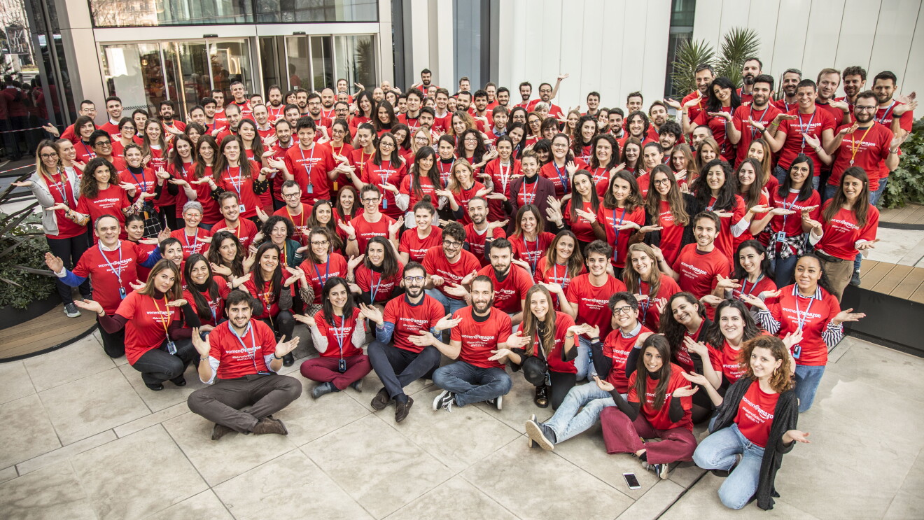 Gruppo di persone in posa con una t-shirt rossa ed entrambe le mani alzate a simulare una bilancia. Posano davanti all'ingresso di un ufficio Amazon.