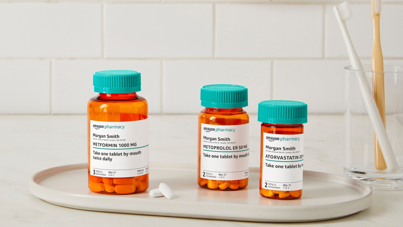 Three Amazon Pharmacy pill bottles sit on a white tray next to a glass with two toohbrushes in it.