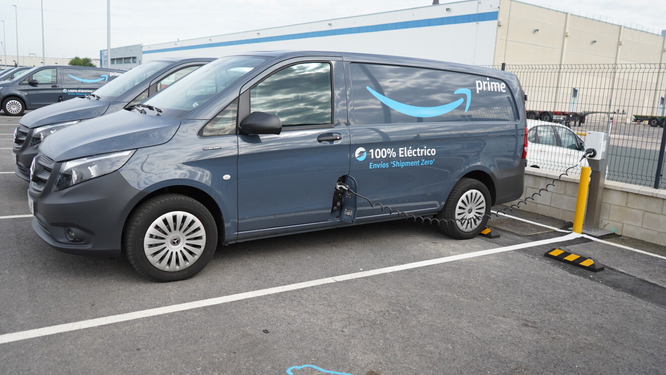 Una Mercedes Benz-Vans en un aparcamiento. La furgoneta es de color negro, con la sonrisa de color azul y la palabra Prime en blanco. Además, la furgoneta lleva la siguiente frase: 100% Eléctrico Envíos 'Shipment Zero'. La furgoneta está conectada a la red eléctrica y se está cargando la bateria. 
