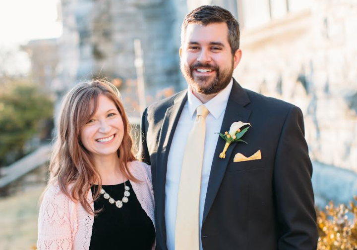 An image of a man and a woman smiling outside of a building while dressed in more formal clothing/