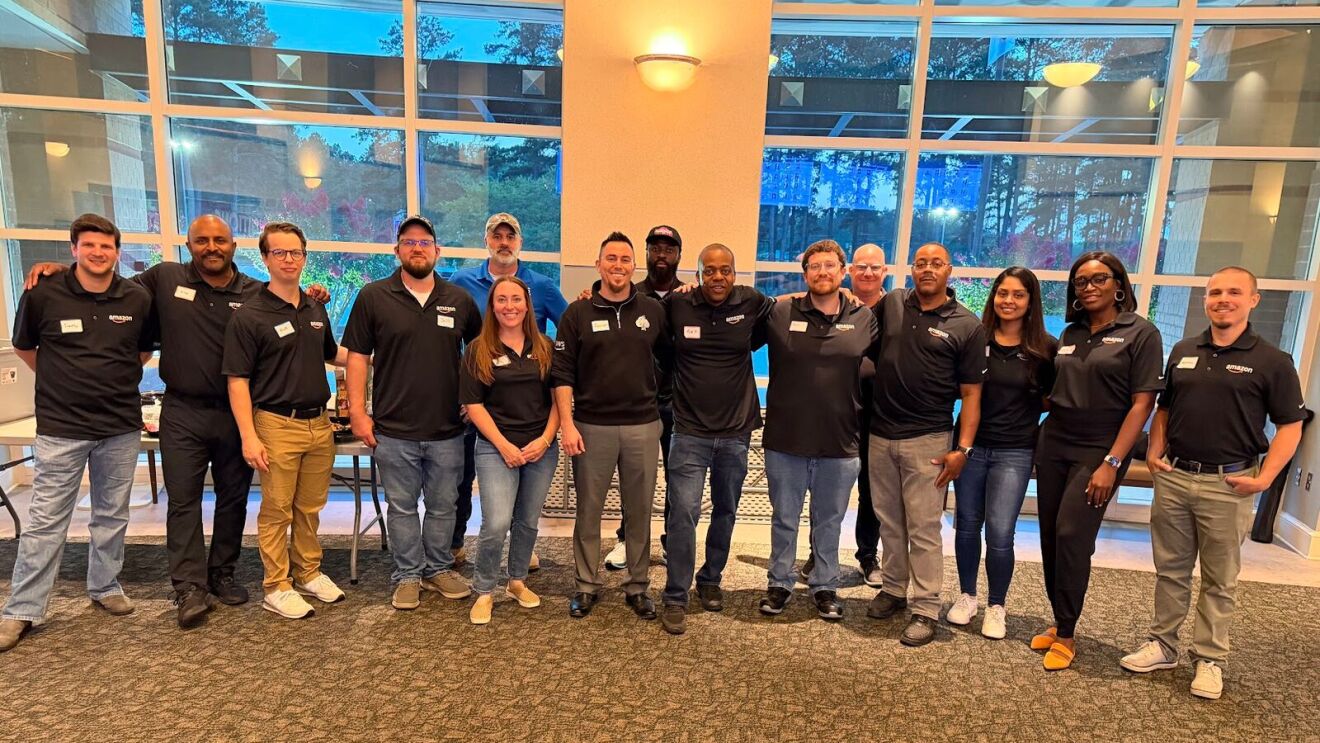 Team of approximately 15 professionals wearing matching black polo shirts standing together in a modern office building with floor-to-ceiling windows