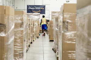 A man wearing shorts, tennis shoes, a t-shirt and safety vest walks between pallets of supplies in Amazon's Disaster Relief Hub