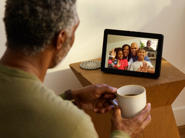 An Echo Show device on a wooden side table. On the screen, a family of four are smiling and waving on the screen. Facing the device is a man, holding a coffee cup. The screen shows his smiling face.