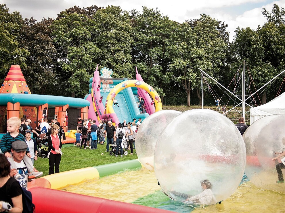 Verschiedene Spielstationen auf dem Spielfest: Man sieht Besucher und riesige Ballon, die auf einem aufblasbaren Schwimmbecken treiben, Im Balloninneren sind Kinder.