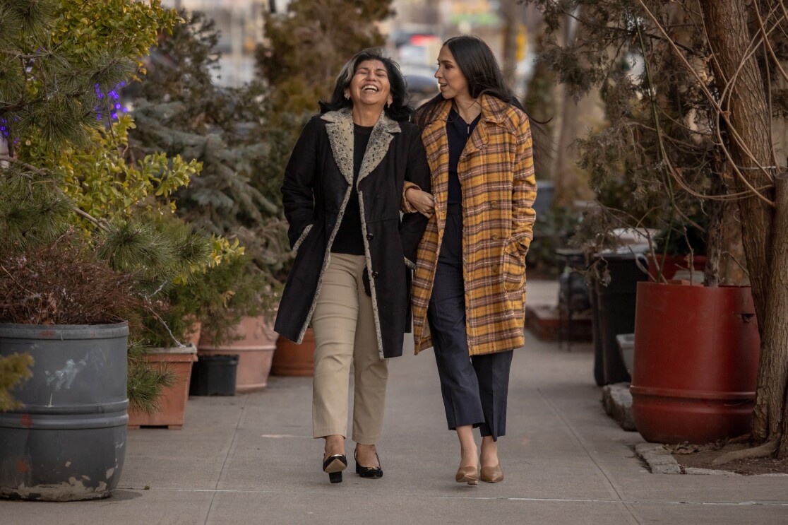 Two women walk arm in arm down a city street flanked by large potted plants and trees.